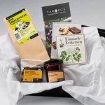 Geschenkkorb mit weißen Seidenpapier-Einlagen und verschiedenen Samocca-Produkten. Enthalten sind ein Beutel Bio-Früchtetee, ein Glas Himbeer-Fruchtaufstrich, Kürbiskern-Chili-Cookies sowie mehrere Informationskarten und Einmach-Etiketten. Die Produkte sind dekorativ in einer schwarzen Geschenkbox angeordnet. Die sommerliche Variante des Korbs enthält keine Schokolade.