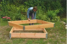 Mann baut im Garten ein Hochbeet aus Holzrahmen zusammen und setzt dabei ein weiteres Rahmenelement auf die untere Konstruktion.